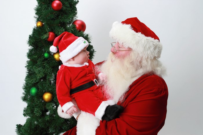 Santa Claus holding a baby in a Santa outfit by a Christmas tree