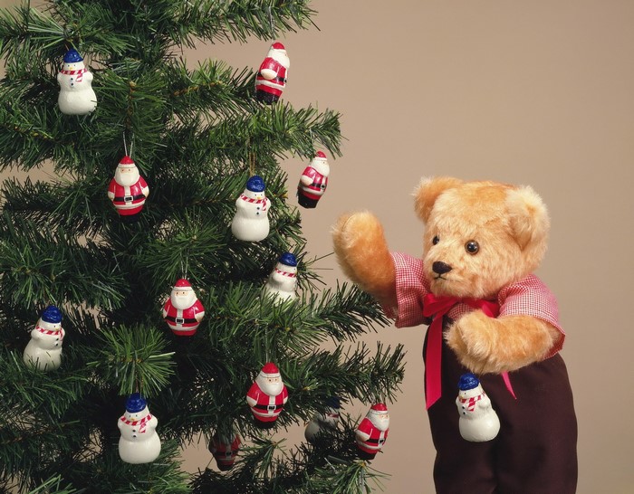 Teddy bear decorating a Christmas tree with Santa and snowman ornaments
