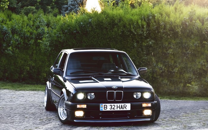 Black BMW E30 parked on cobblestone with green hedge background