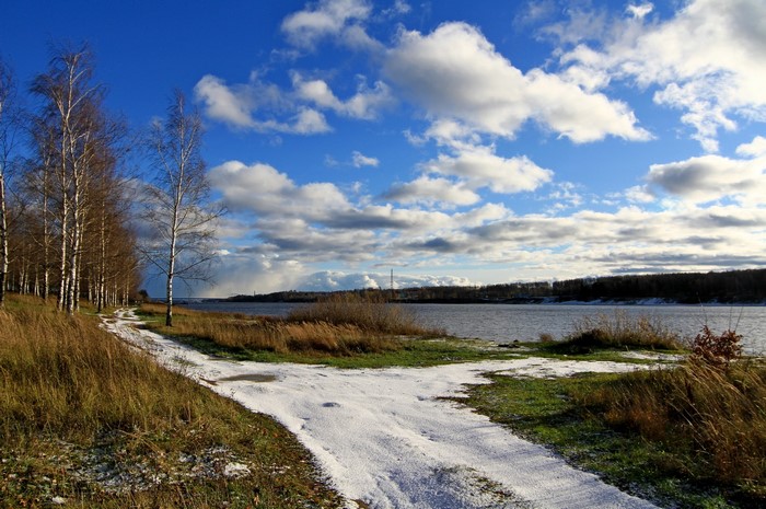 Snowy path by a river with birch trees under a cloudy sky