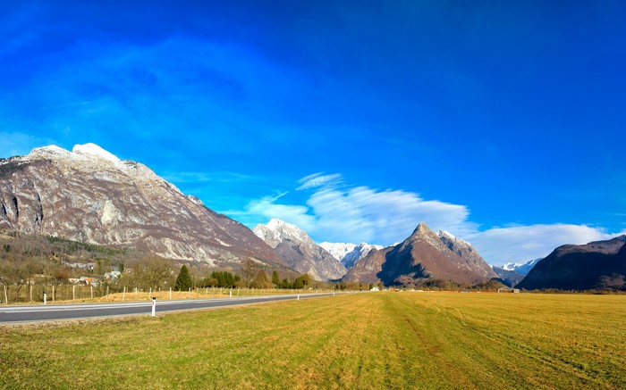 Road through a grassy field with mountains under a blue sky