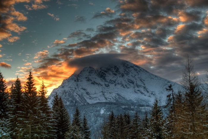 Snowy mountain peak with dramatic sunset clouds and pine forest