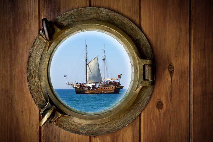 Wooden porthole looking out at a sailing ship on the sea