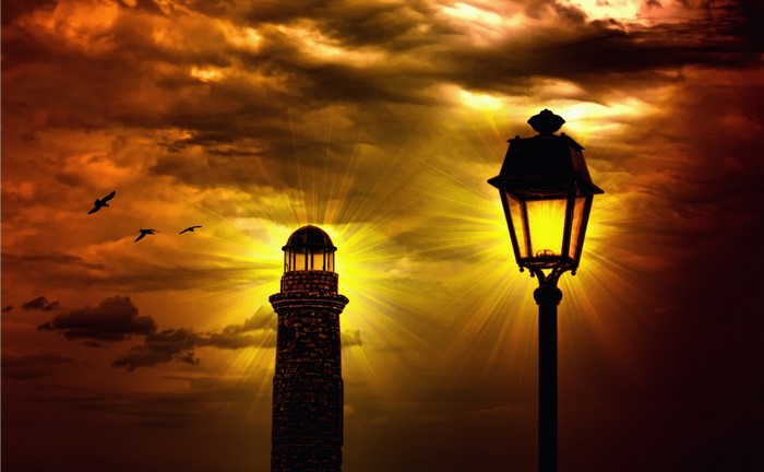 Lighthouse and street lamp against a dramatic sunset sky with birds