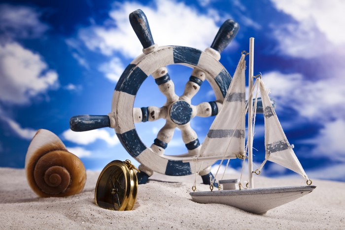 Toy sailboat, ship's wheel, shell, and compass on sand