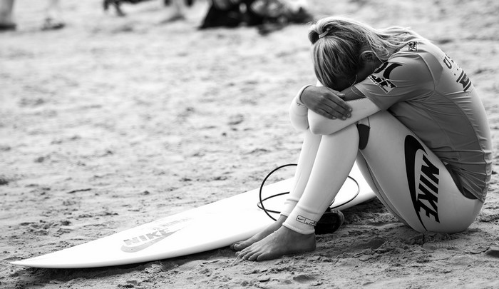Surfer in Nike wetsuit sitting on beach next to surfboard