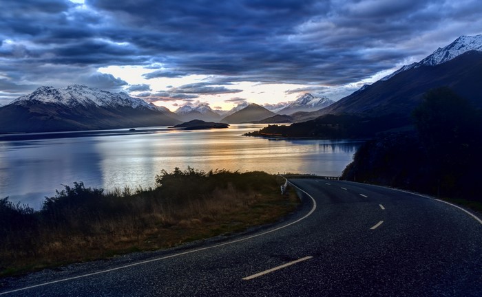 Road by a lake with snow-capped mountains and clouds