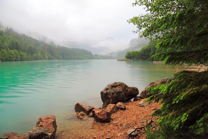 Turquoise lake surrounded by misty, forested mountains and rocky shore