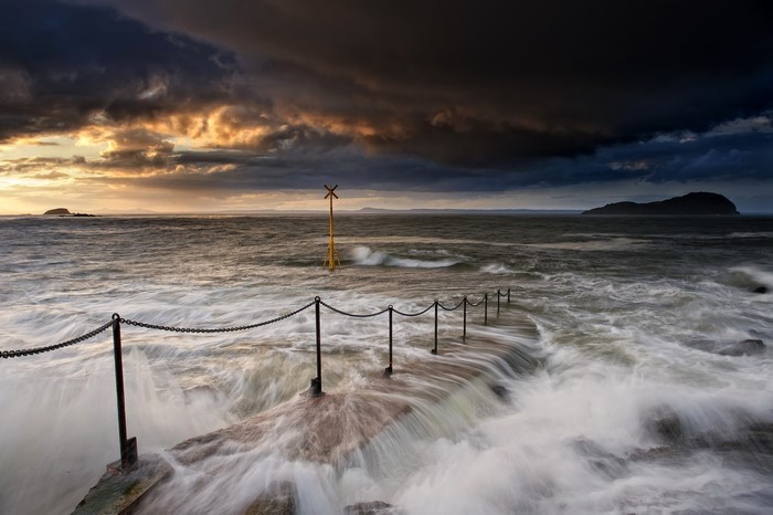 Waves crashing on a pier with a yellow marker in North Berwick, Scotland