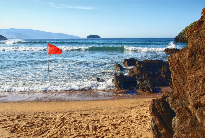 Red flag on a sandy beach with waves and rocks