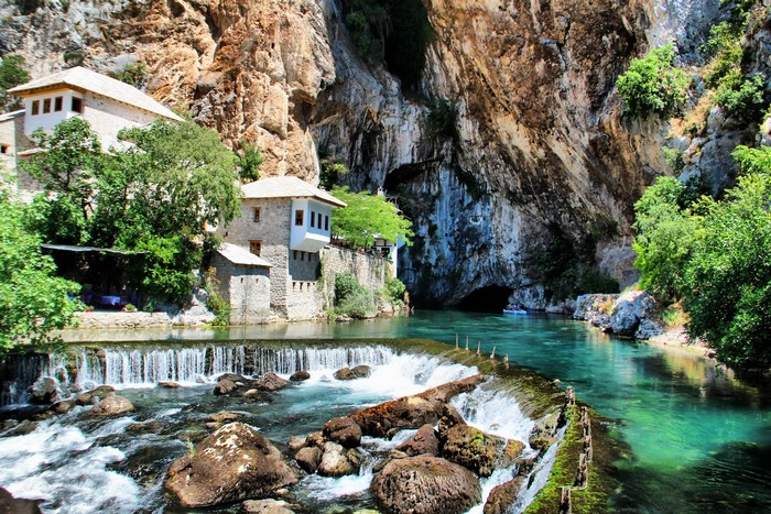 Stone buildings by a turquoise river and waterfall