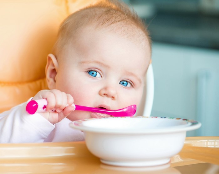 Baby with blue eyes holding a pink spoon near a bowl