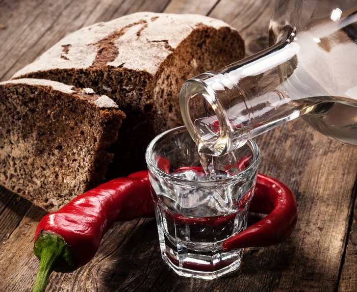 Vodka being poured into a shot glass with chili peppers and bread