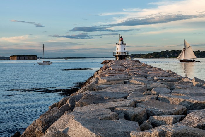 Lighthouse on a stone jetty with sailboats and a fort