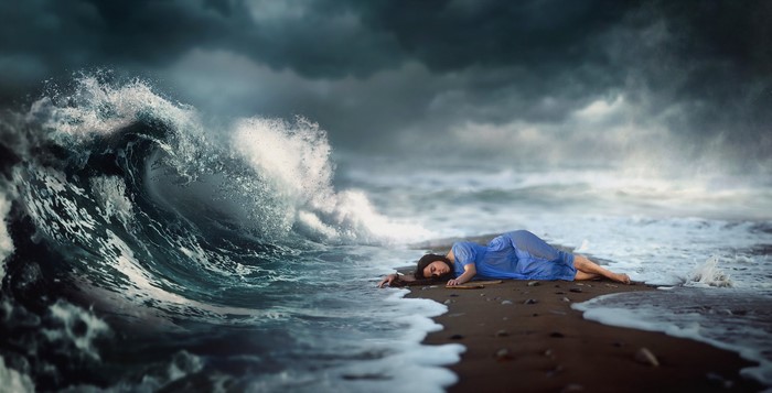 Woman in blue dress lying on beach with large wave