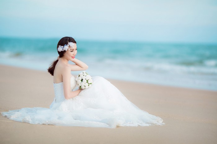 Bride in Wedding Dress Sitting on Beach with Bouquet