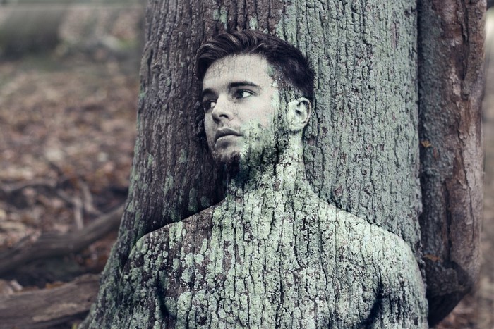 Man with tree bark texture on skin leaning against a tree