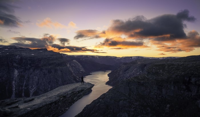 Person standing on Trolltunga cliff overlooking a fjord at sunset