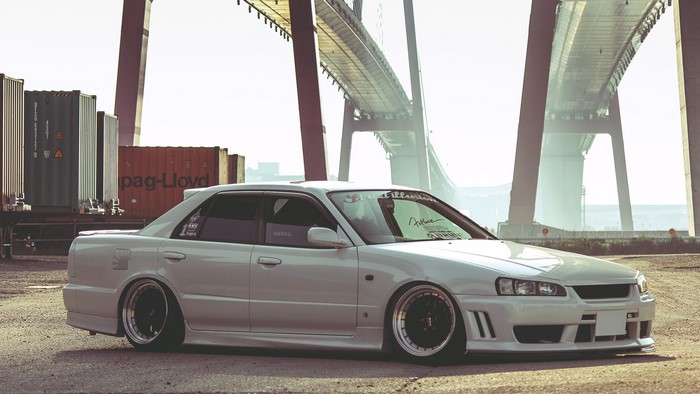 White Nissan Skyline sedan parked near shipping containers and a bridge