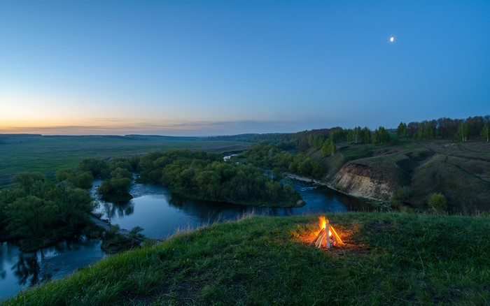 Campfire by a river at dusk with moon