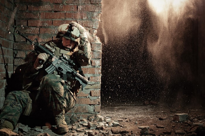 Soldier in tactical gear with assault rifle behind brick wall