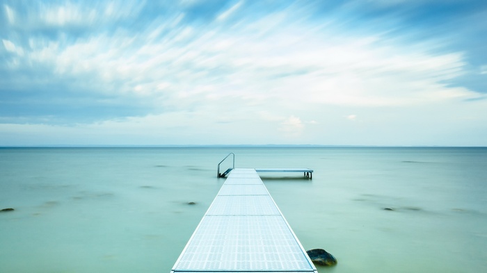 A long pier extends into a calm sea under a cloudy sky.