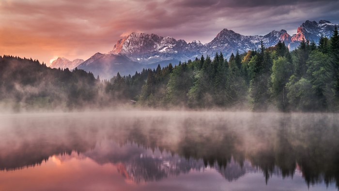 Misty lake with forest and snow-capped mountains at sunrise