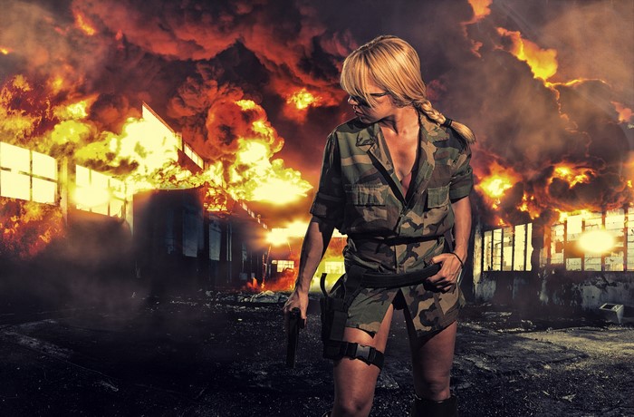 Blonde woman in camouflage uniform with gun in front of fire