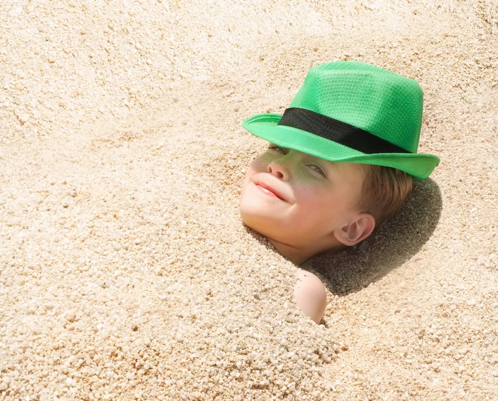 Boy buried in sand wearing a green hat