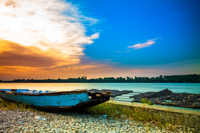 Old blue boat on a rocky shore by the river at sunset