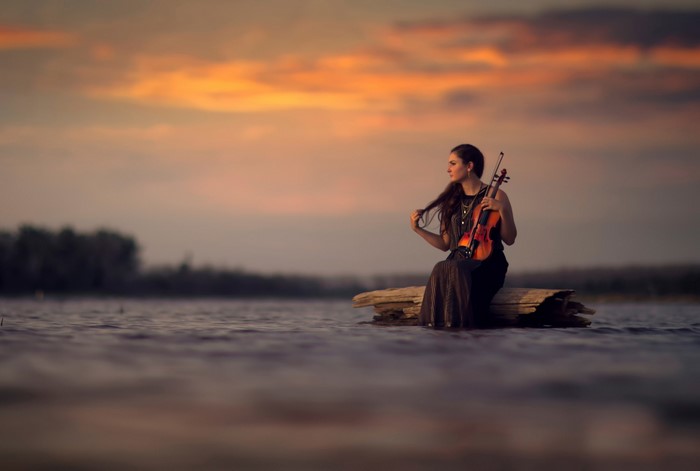 Woman with violin sitting on a log in water at sunset