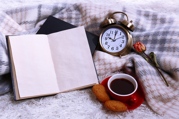 Alarm clock, open book, coffee, cookies, and rose on blanket