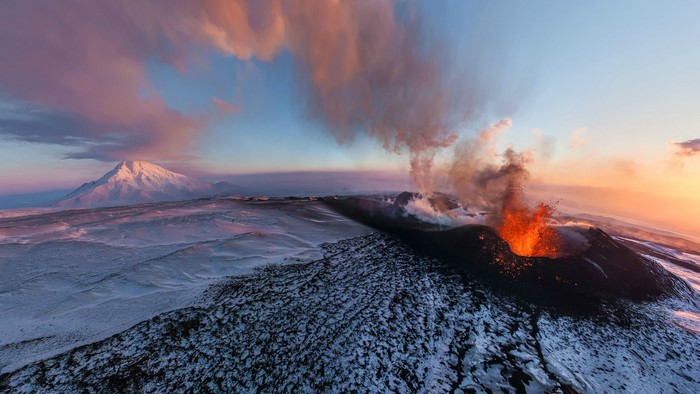 Volcano erupting at sunset with snow-covered mountains