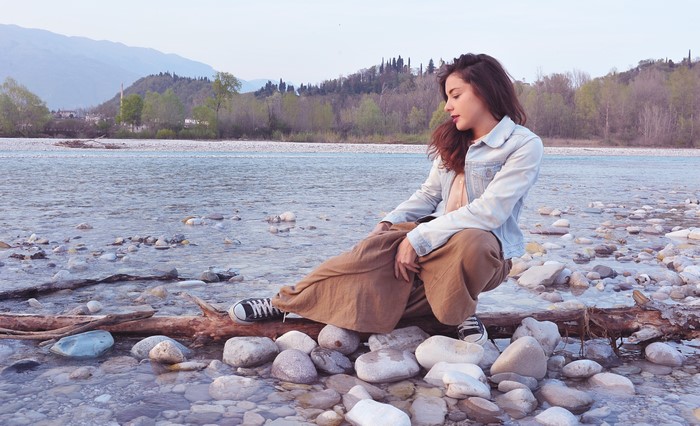Woman sitting by a river with mountains in the background