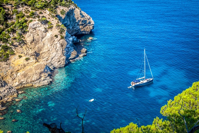 Sailboat and small boat near rocky cove with trees