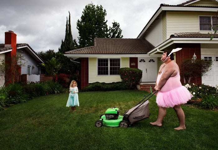 Man in tutu mowing lawn with girl watching