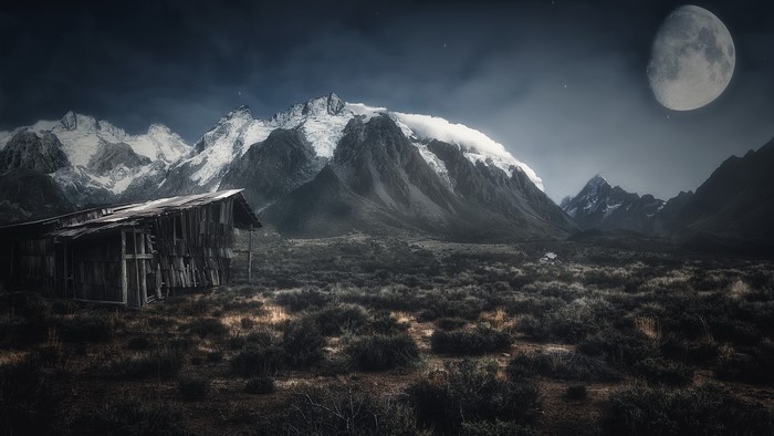 Abandoned cabin in front of snowy mountains under the moon