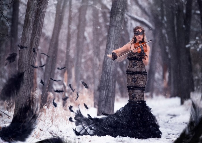 Woman in black dress playing violin in snowy forest with feathers