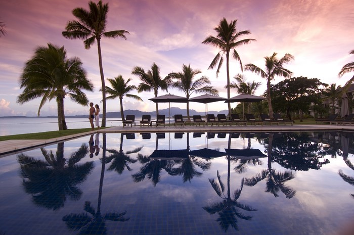 Couple by pool with palm trees and ocean at sunset