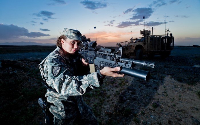 Soldier aiming a rifle with military vehicle in background