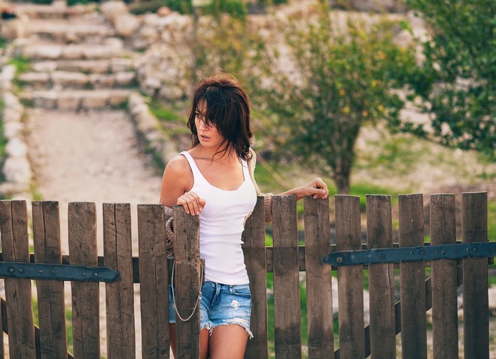 Woman in white singlet and denim shorts leaning on a wooden fence