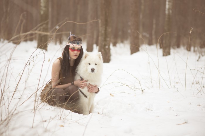 Woman in Native American attire with Samoyed dog in winter forest