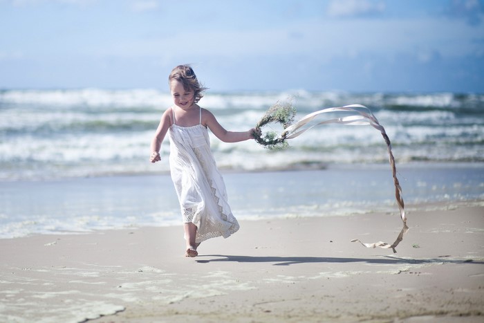 Little girl in white dress running on beach with wreath