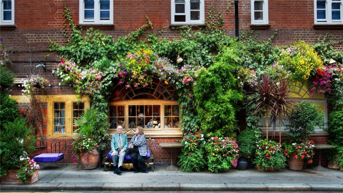 Two people sitting on a bench in front of a brick building covered in plants and flowers
