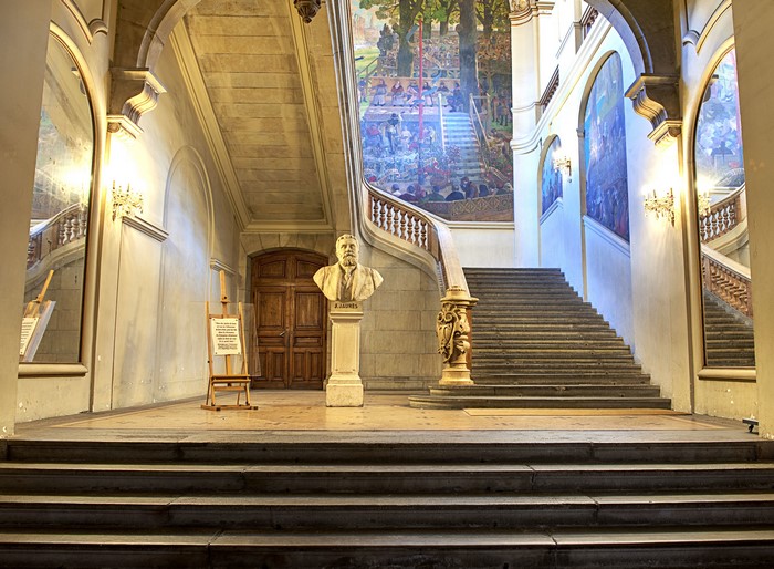 Bust of a man on a pedestal in front of a grand staircase with murals