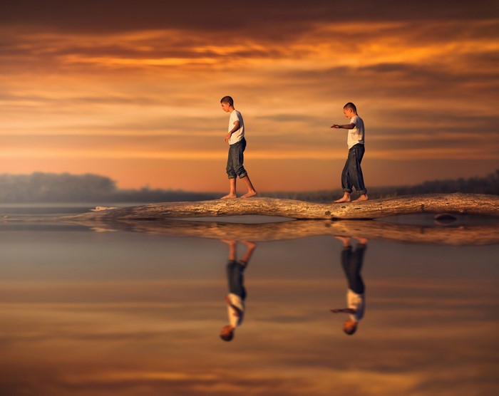 Two boys walking on a log over water at sunset