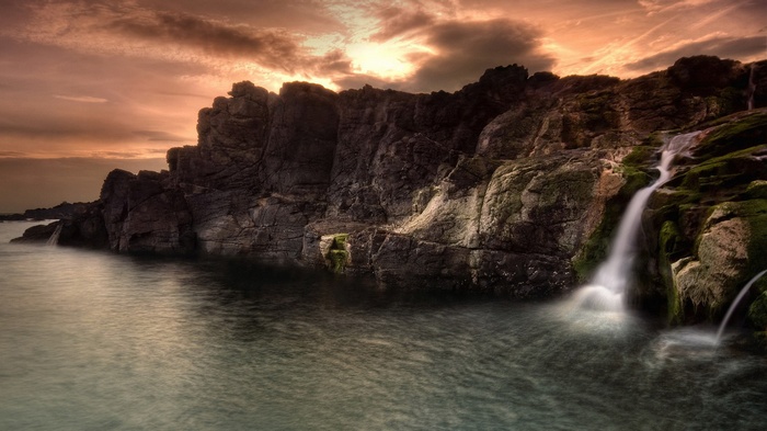 Waterfall cascading over rocky cliffs into the ocean at sunset