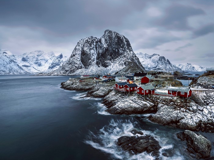 Red fishing cabins on rocky shore with snow-capped mountains