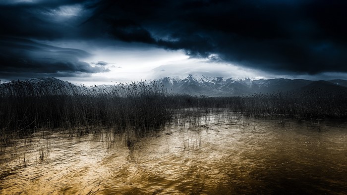 Lake with reeds and snow-capped mountains under thunderclouds