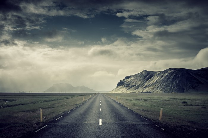 Empty road through a vast landscape with mountains under a cloudy sky